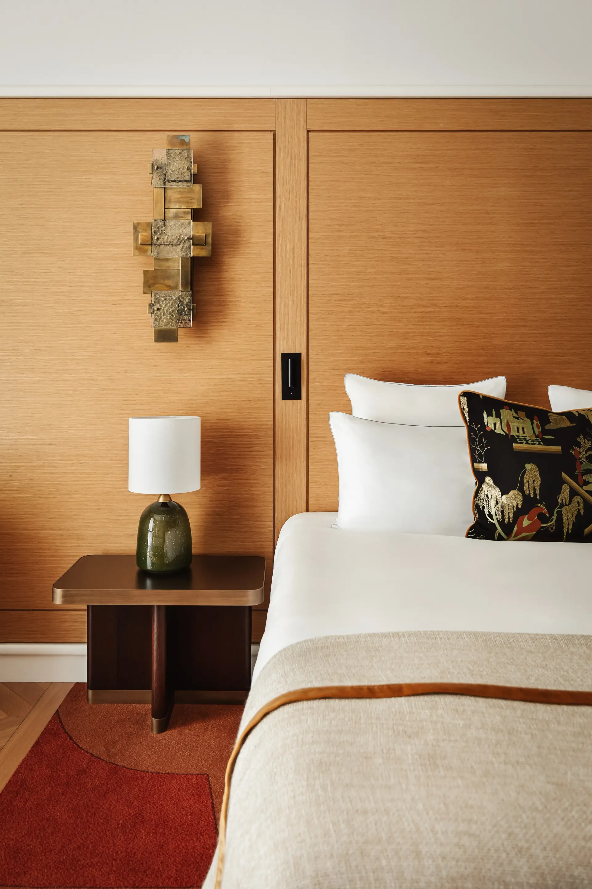 Partial view of an elegant hotel room featuring a comfortable bed with white linens, a beige bedspread and a black cushion adorned with golden and colorful Asian patterns. The headboard wall is covered in warm light wood panels, enhanced by a sculptural gold metal wall sconce. Next to the bed, a dark nightstand holds a small table lamp with a green glass base and white shade, all on a rug in terracotta and red tones.