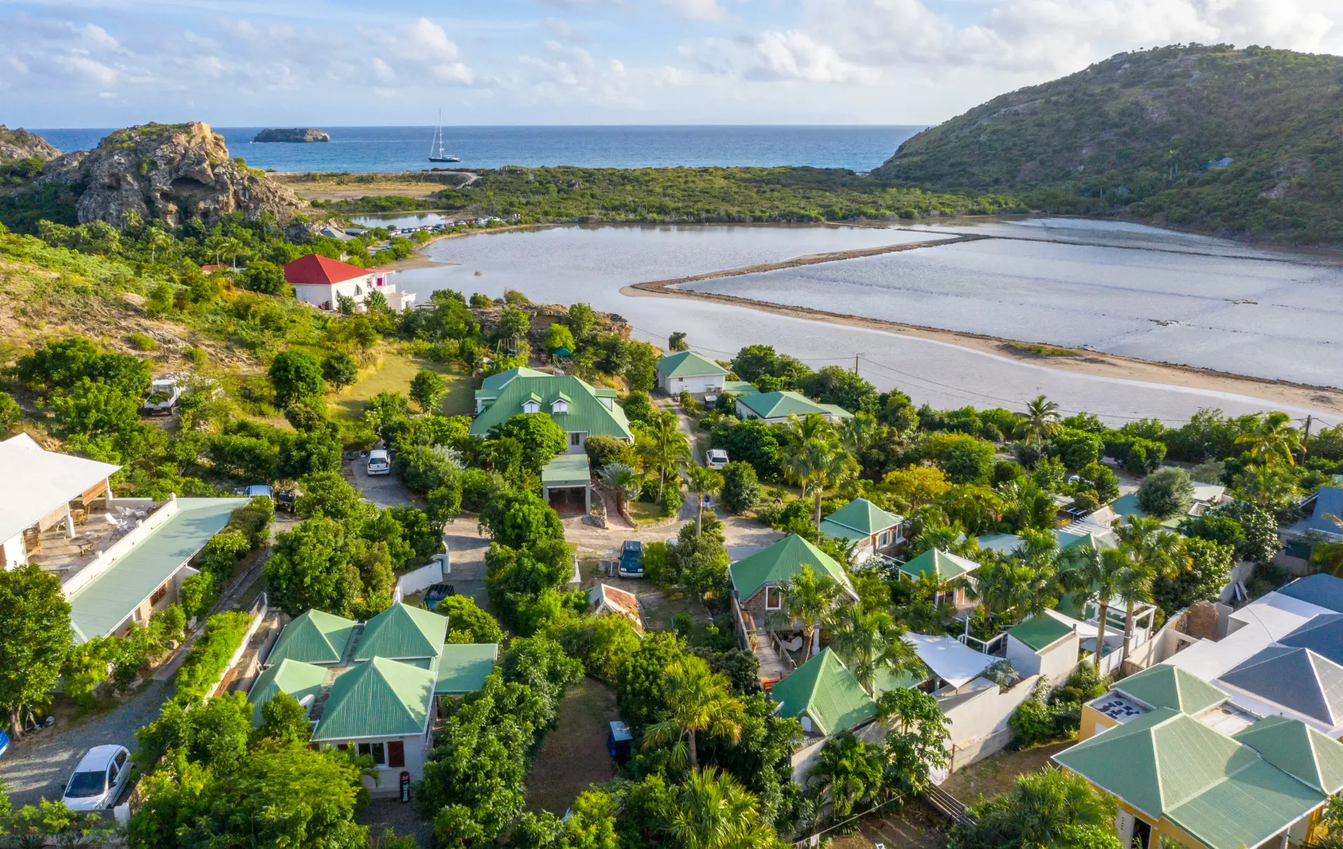 Vue aérienne panoramique d'un complexe résidentiel composé de plusieurs villas aux toits verts, nichées dans une végétation tropicale luxuriante près d'un étang salin et de la mer. Le paysage est caractérisé par un relief vallonné, un littoral côtier et une eau turquoise en arrière-plan sous un ciel partiellement nuageux, offrant une perspective globale sur ce lieu de villégiature.