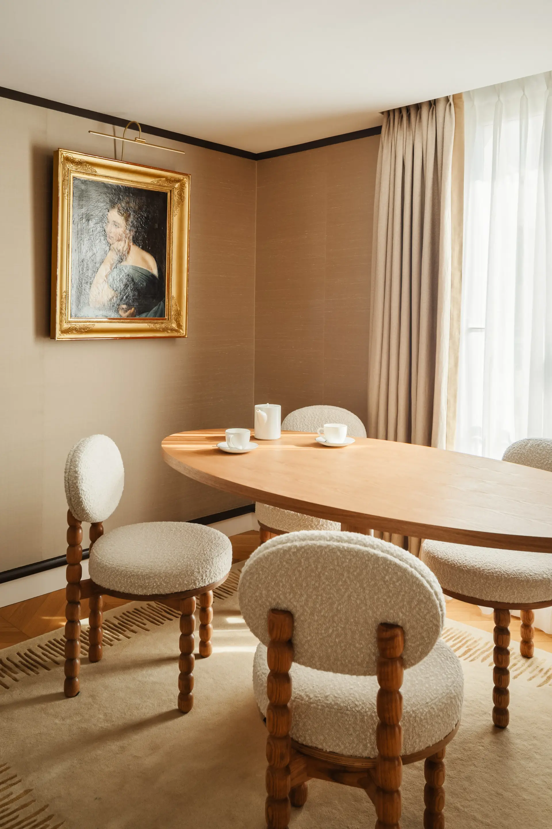 Elegant dining room with a light wood oval table surrounded by chairs with an original design, featuring turned wooden legs and rounded seats and backs covered in textured cream fabric. On the beige wall, a classic portrait in a gold frame is highlighted by a picture light, and the room is completed by a matching rug and light curtains falling in front of a window.