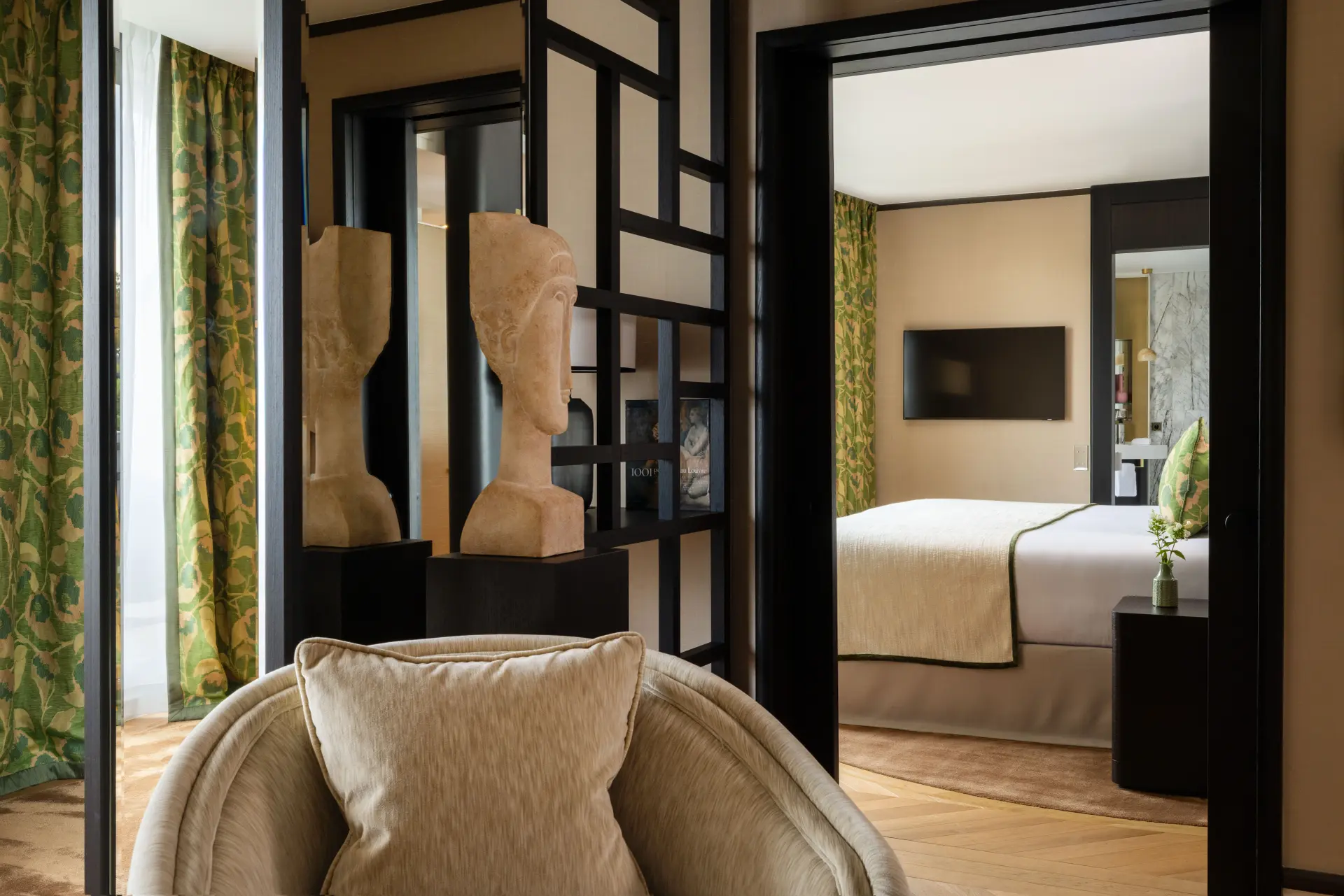 Elegant interior view of a suite, highlighting a rounded cream armchair with a matching cushion in the foreground. In the center, a dark wood openwork partition separates the spaces and displays a stylized bust sculpture. Through a door, a bright bedroom with a comfortable bed and wall-mounted TV can be seen, all enhanced by green botanical-patterned curtains that add a natural touch.