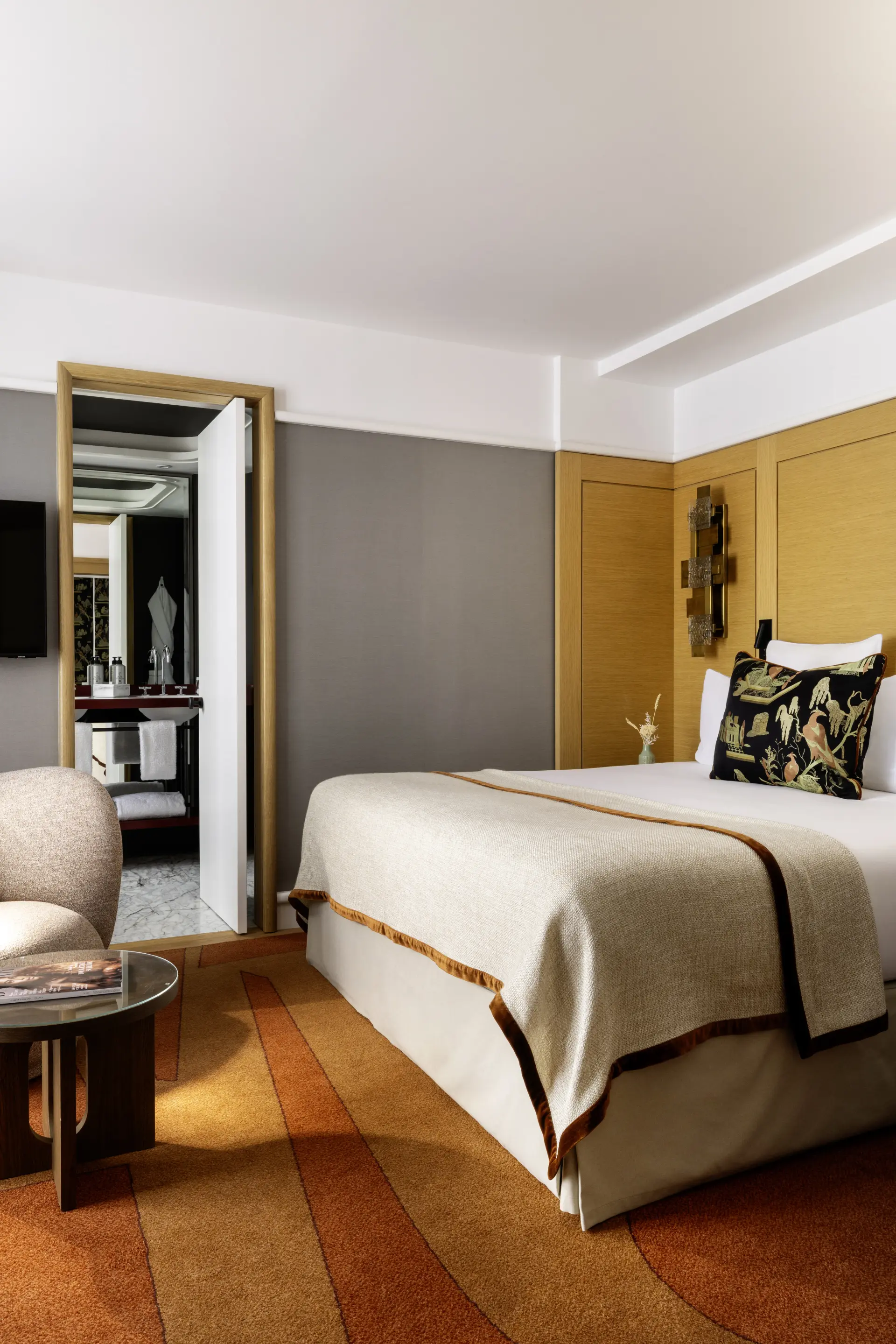 Interior view of an elegant, modern hotel room, highlighting a large comfortable bed adorned with a beige bedspread and a decorative black cushion with patterns. On the left, an open door reveals a bright bathroom with a vanity and mirror. The floor is covered in warm orange and brown carpet, and a small seating area with an armchair and coffee table is visible in the foreground, creating a welcoming, refined atmosphere.